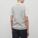 A model with dark brown hair wears the sleeveless knit top in grey melange with navy trousers show backside against a white backdrop.