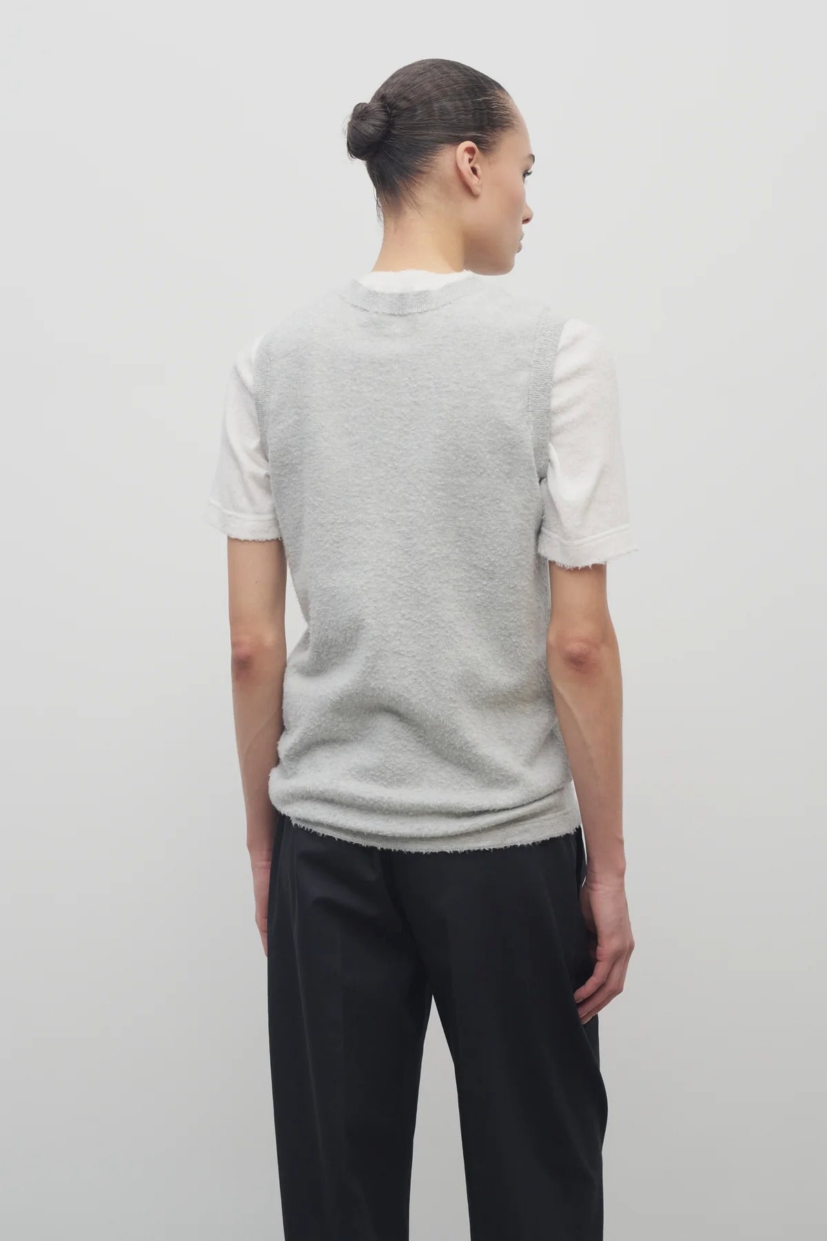 A model with dark brown hair wears the sleeveless knit top in grey melange with navy trousers show backside against a white backdrop.