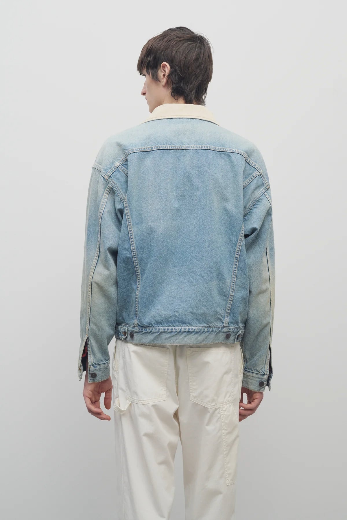 Back view of light indigo collared joret jacket on model wearing white trousers, against a white background.