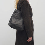 Front view of black lilou woven tote on model wearing brown overcoat against a white backround.