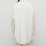 Back view of white luka shirt on model wearing white pants, against a whtie background.
