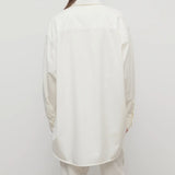 Back view of white luka shirt on model wearing white pants, against a whtie background.