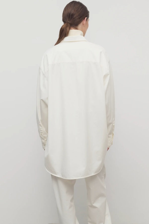 Back view of white luka shirt on model wearing white pants, against a whtie background.