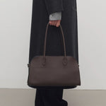 Front view of deep brown margaux shoulder 12 bag on model wearing grey overcoat, green shirt, black pants and mules, against a white background.