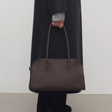 Front view of deep brown margaux shoulder 12 bag on model wearing grey overcoat, green shirt, black pants and mules, against a white background.