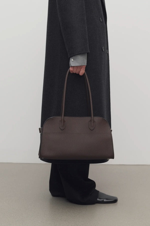 Front view of deep brown margaux shoulder 12 bag on model wearing grey overcoat, green shirt, black pants and mules, against a white background.