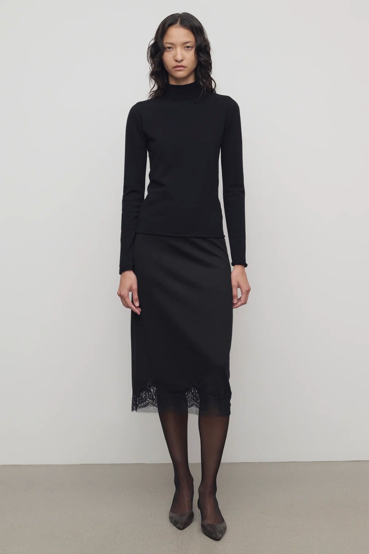 Front view of black mea skirt on model wearing black top and heels, against a white background.