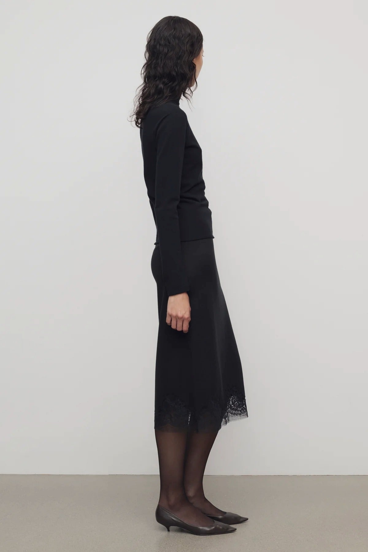 Side view of black mea skirt on model wearing black top and heels, against a white background.