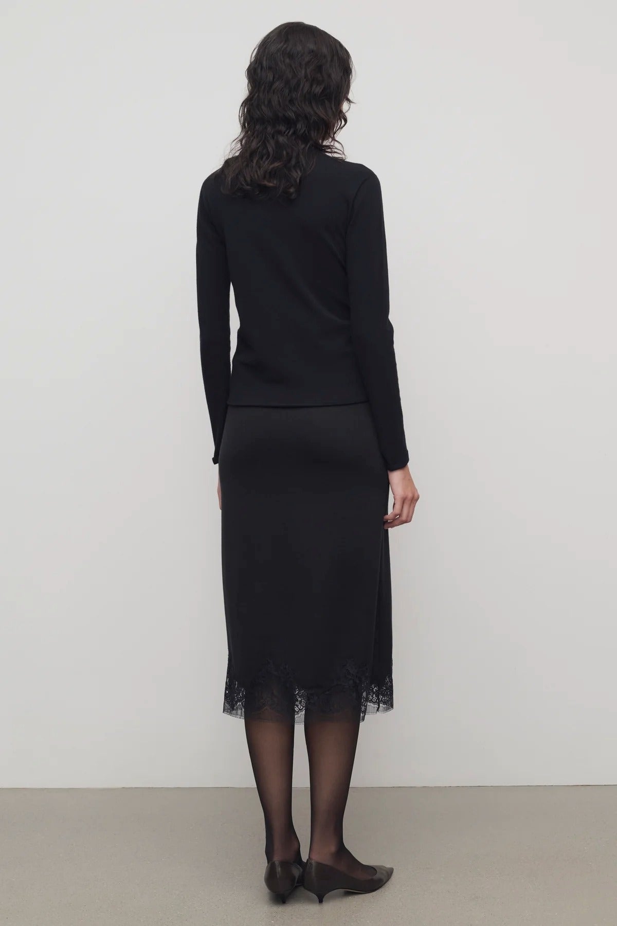 Back view of black mea skirt on model wearing black top and heels, against a white background.