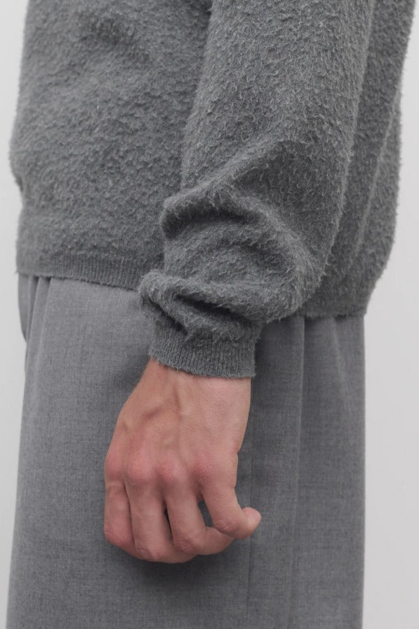 Side closeup view of grey melange almot sweater on model wearing grey pants, against a white background.