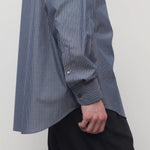 Closeup side view of blue/white men's miller stripe shirt on model wearing black trousers, against a white background.