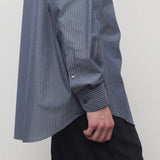 Closeup side view of blue/white men's miller stripe shirt on model wearing black trousers, against a white background.