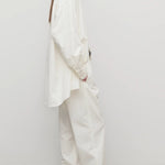 Side view of white neriah pants on model wearing ivory top, jacket and sneakers, against qa white background.