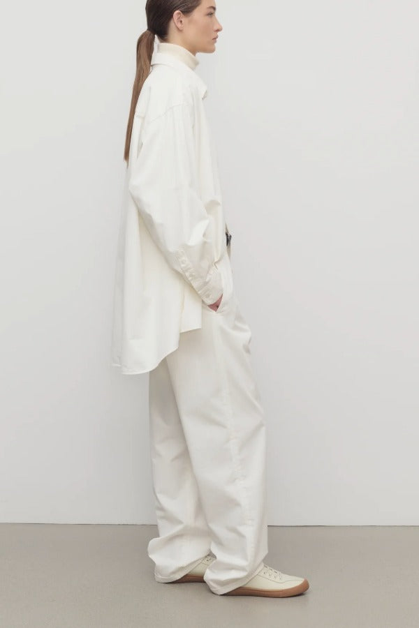 Side view of white neriah pants on model wearing ivory top, jacket and sneakers, against qa white background.