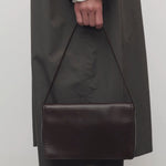 Front view of dark brown nuance bag on model wearing grey trench coat and khaki pants, against a white background.