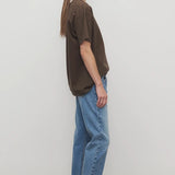 Side view of indigo riaco jean on model wearing brown shirt and sneakers, against a white background.