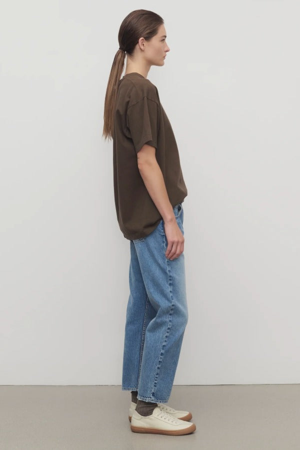 Side view of indigo riaco jean on model wearing brown shirt and sneakers, against a white background.