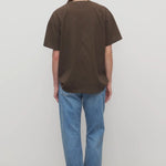 Back view of indigo riaco jean on model wearing brown shirt and sneakers, against a white bakground.
