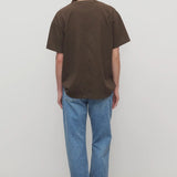Back view of indigo riaco jean on model wearing brown shirt and sneakers, against a white bakground.