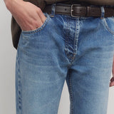 Front closeup view of indigo riaco jean on model wearing brown shirt and belt, against a white background.