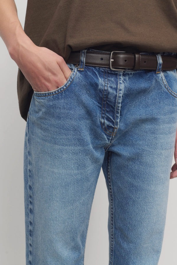 Front closeup view of indigo riaco jean on model wearing brown shirt and belt, against a white background.