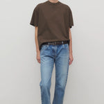 Front view of medium blue riaco jean on model wearing brown shirt and white sneakers, against a white background.