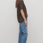 Side view of medium blue riaco jean on model wearing brown shirt and white sneakers, against a white background.