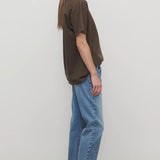 Side view of medium blue riaco jean on model wearing brown shirt and white sneakers, against a white background.