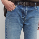 Front closeup view of medium blue riaco jean on model wearing brown shirt, against a white background.
