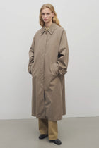 A model with long blonde hair wears a loose-fitting, taupe trench coat over wide-legged tan trousers and black shoes, standing against a plain white background.