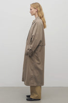 Long blonde model at side angle wears long taupe trench coat with khaki trousers and black loafers against a white background.