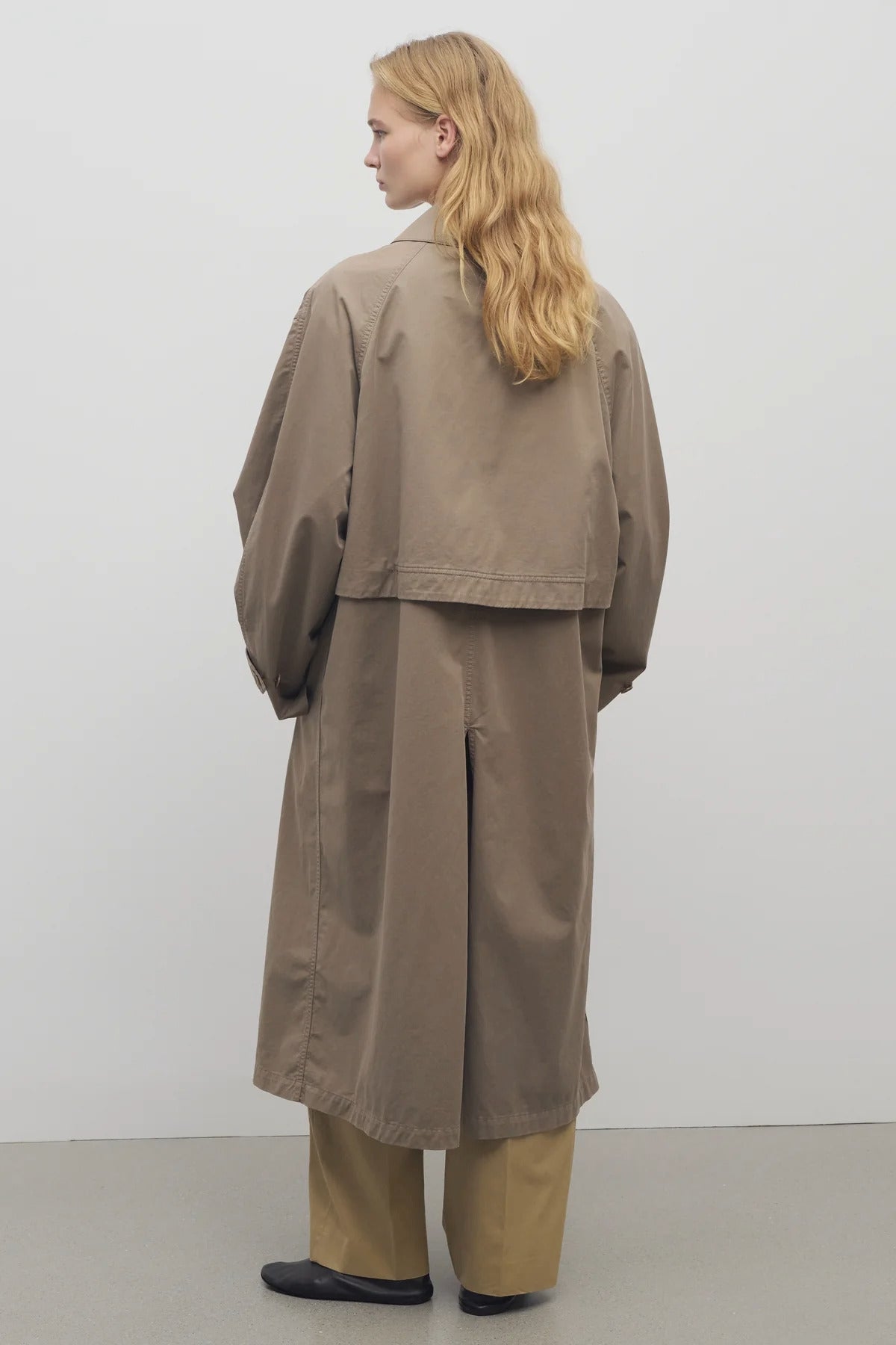 A model with long blonde hair at a backside viewpoint wears a taupe long trench coat with khaki trousers and black loafers against a white background.