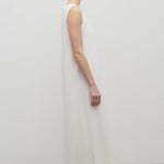Side view of light-brown-haired model in sleeveless round neck maxi white cotton dress and sandals on plain white background
