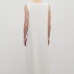 Back view of light-brown-haired model wearing sleeveless round neck maxi white cotton dress and sandals on plain white background.