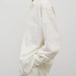 Side view of white erika shirt on model wearing white trousers, against white background.