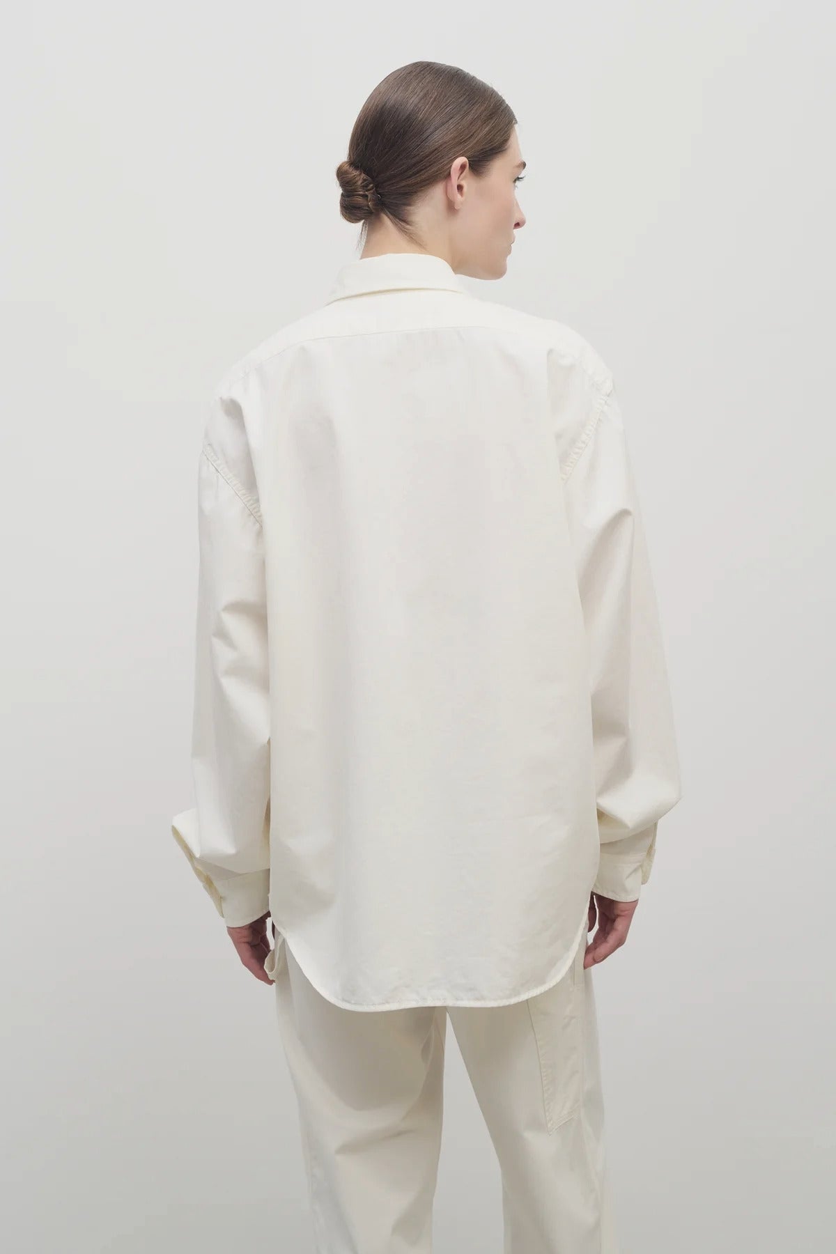 Back view of white erika shirt on model wearing white trousers, against white background.