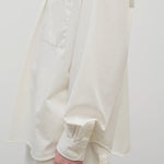 Side closeup view of white erika shirt on model wearing white trousers, against white background.