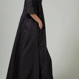 Side view of black rachel long dress on model wearing black flats, against a white background.