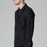 Side view of black regular 2 button jacket on model wearing black top and pants, against a grey background.