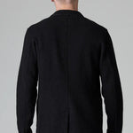 Back view of black/brown regular fit jacket on model wearing black pants, against a grey background.