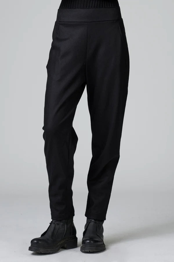 Front view of black slim fit trousers on model wearing black top and boots, against a grey background.