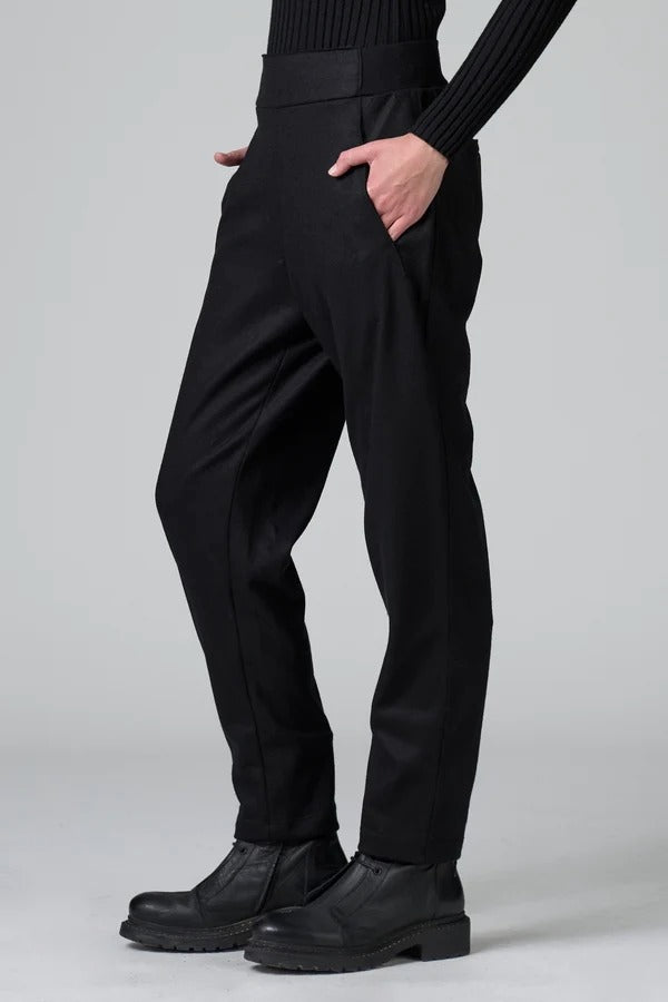 Side view of black slim fit trousers on model wearing black top and boots, against a grey background.