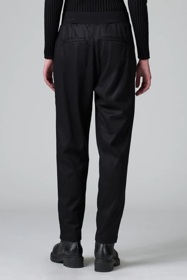 Back view of black slim fit trousers on model wearing black top and boots, against a grey background.