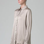 Side view of stone stretch silk satin regular fit shirt on model wearing chalk pants, against a grey background.