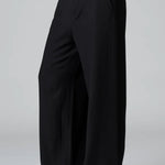 Side view of black stretch wide leg trousers on model wearing black top and boots, against a grey background.