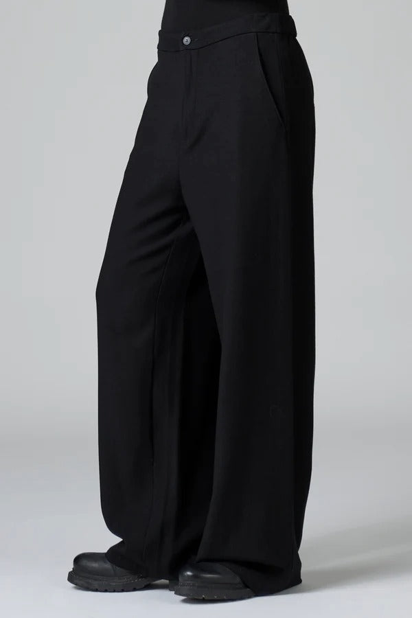 Side view of black stretch wide leg trousers on model wearing black top and boots, against a grey background.