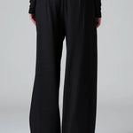 Back view of black stretch wide leg trousers on model wearing black top and boots, against a grey background.