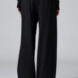 Back view of black stretch wide leg trousers on model wearing black top and boots, against a grey background.