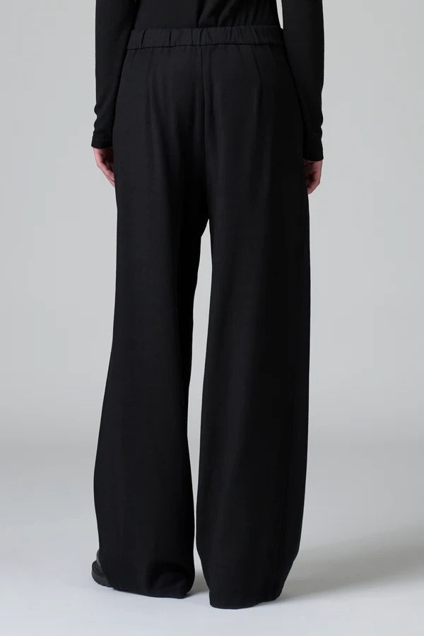Back view of black stretch wide leg trousers on model wearing black top and boots, against a grey background.