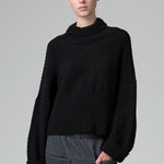 Front view of black wool chain stitch knit ribbed turtleneck on model wearing grey pants, against a grey background.